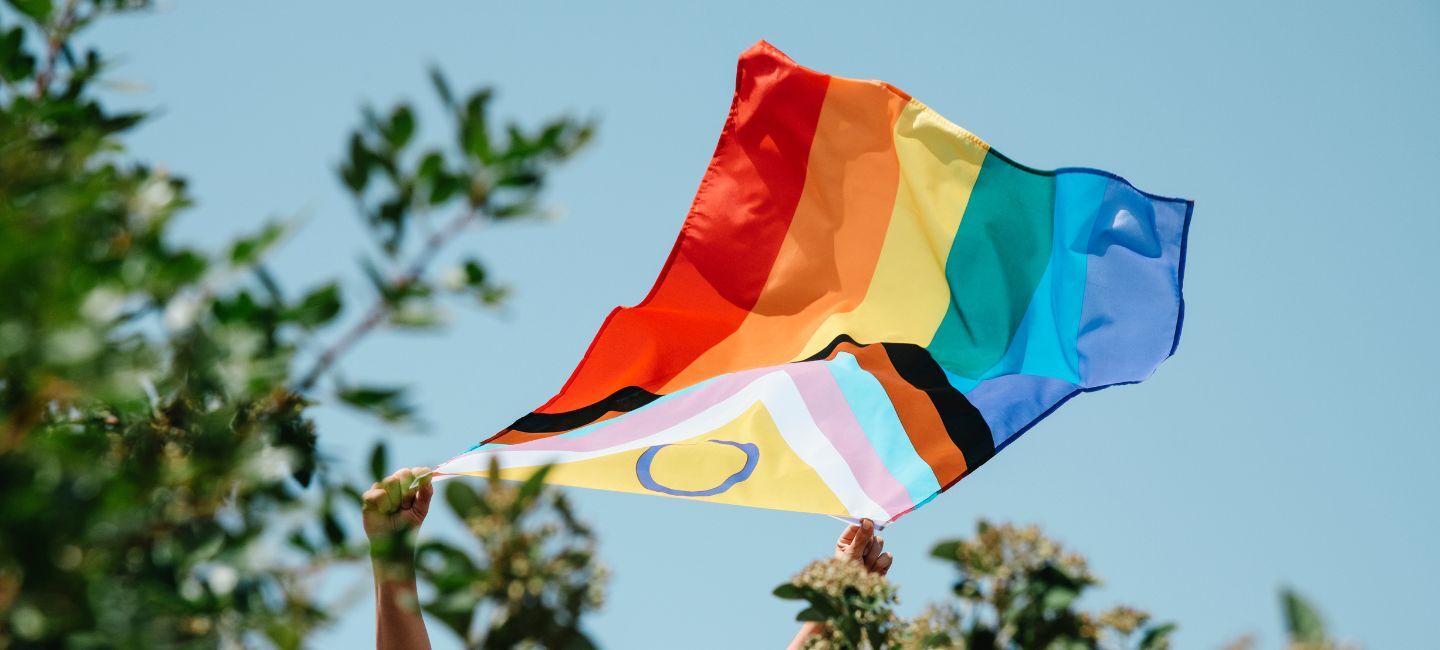 An inclusive Pride flag is help up against a blue sky.